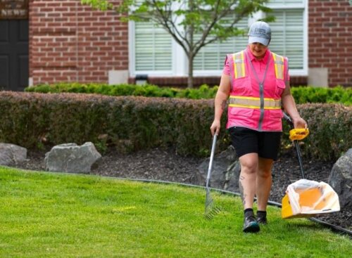Pet waste removal technician in a hi-vis vest carrying a rake and bag across a residential lawn